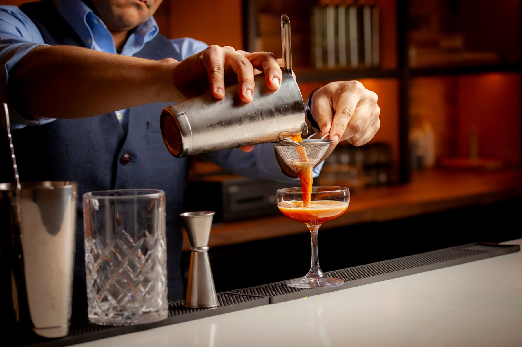 Hands pouring a cocktail through a fine strainer into a coupe at a dimly lit bar