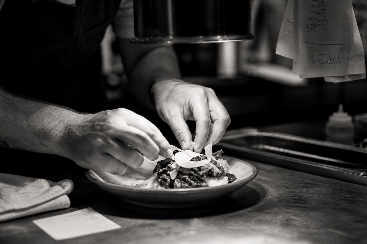 A chef's hands plating a dish at the pass of a small restaurant kitchen, kitchen tickets hanging from the rail. Black and white.